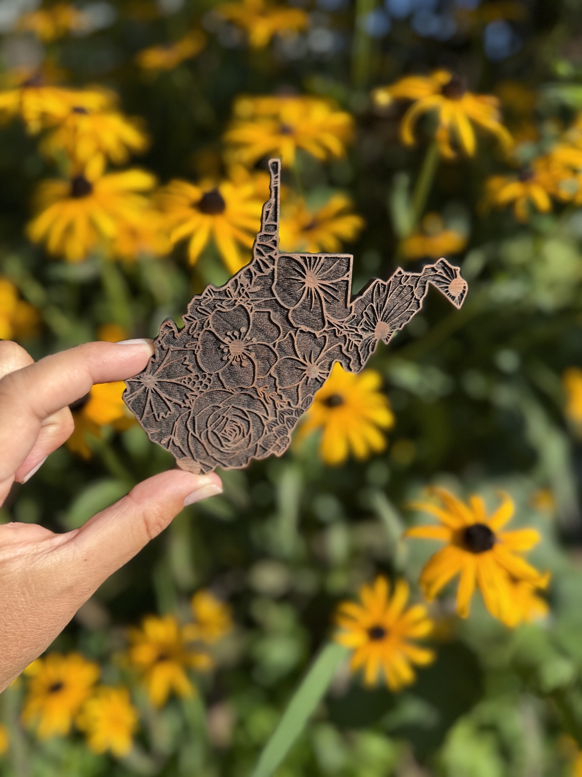 A hand holding a floral engraved West Virginia wood piece in front of yellow flowers